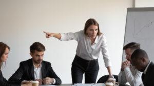 Wütende Chefin schreit traurig gestressten, männlichen Praktikanten bei Teambürotreffen an, Symbol für Diskriminierung bei der Arbeit, Stress und Belästigung am Arbeitsplatz.
